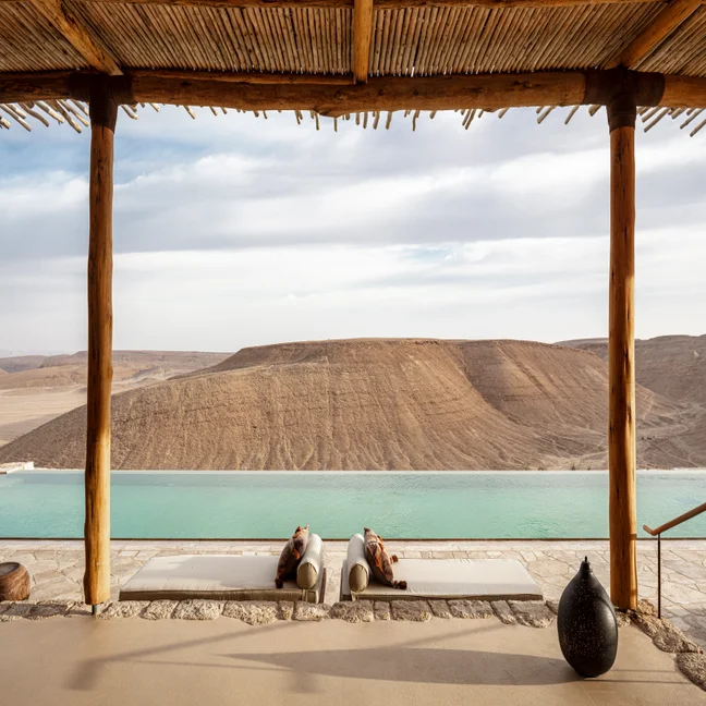 Lounge chairs sit beside an infinity pool under a wooden pergola, offering views of the serene desert and rugged rocky hills.