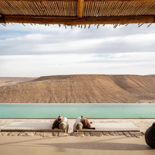 Lounge chairs sit beside an infinity pool under a wooden pergola, offering views of the serene desert and rugged rocky hills.