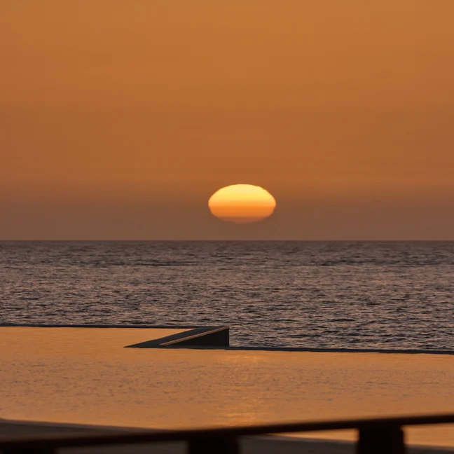 Orange sunlight filters over a tranquil ocean, its reflection visible on the water. An angled structure appears in the foreground against the evening sky.