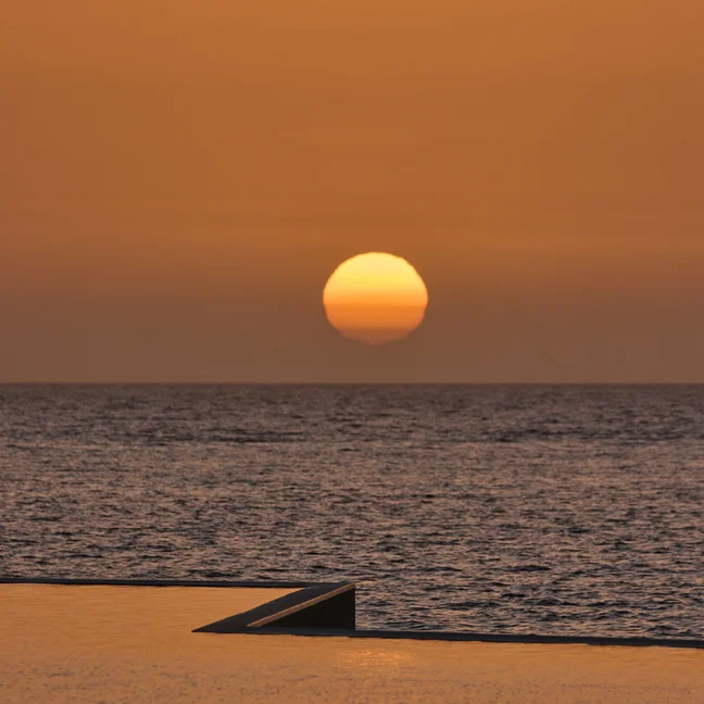 Orange sunlight filters over a tranquil ocean, its reflection visible on the water. An angled structure appears in the foreground against the evening sky.
