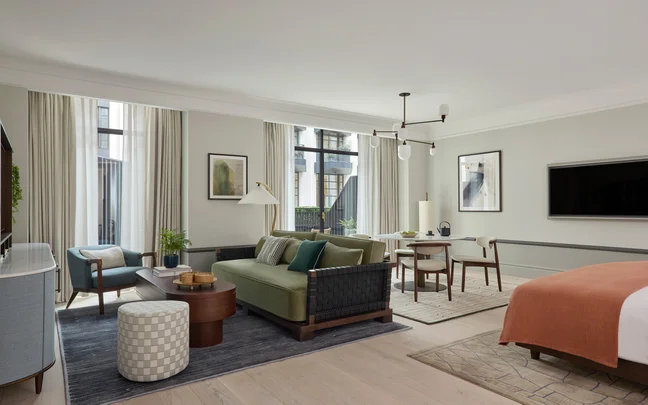 Une suite avec un canapé vert, une table basse, une télévision, une table à manger ronde, un grand lit décoré d'une couverture orange et de grandes fenêtres entourées de rideaux pour la lumière naturelle.