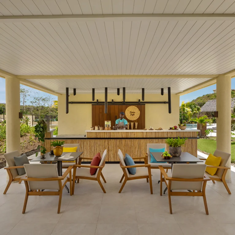 منطقة بار مع طاولات خشبية وكراسي مبطنة ووسائد ملونة؛ نادل على المنضدة. مساحات خضراء مورقة وسماء زرقاء يمكن رؤيتها من خلال المحيط المفتوح.