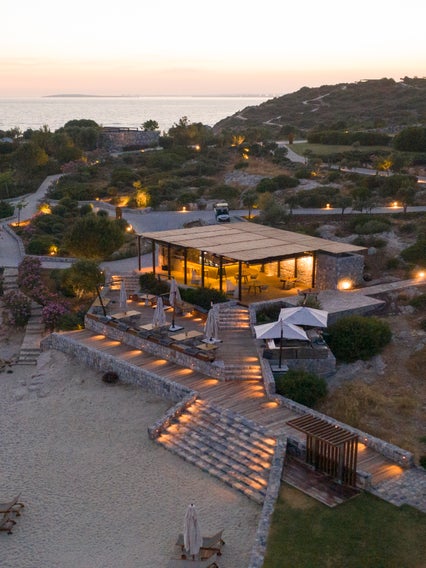 Beachside restaurant with outdoor seating, native greenery, and gentle path lighting, offering views of the sea at dusk. Accessible pathways support ease of movement.