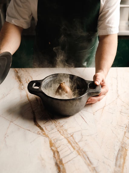 A chef in a dark apron opens a black pot on a marble counter, unveiling freshly prepared food with visible steam rising gently.