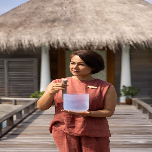 A person in a sleeveless rust outfit stands on a wooden walkway, holding a frosted bowl and mallet, near a thatched-roof building.