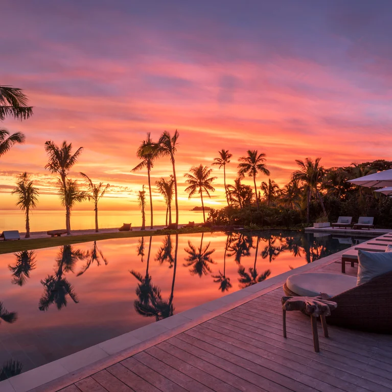 Accessible pool area lined with palm trees, sun loungers, and umbrellas; gentle orange and pink sunset hues reflected on the calm water surface.