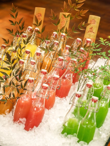 Des jus assortis dans les tons jaune, orange, rose et vert sont placés dans des bacs à glace avec des herbes feuillues et des étiquettes en bois sur une table en bois naturel.