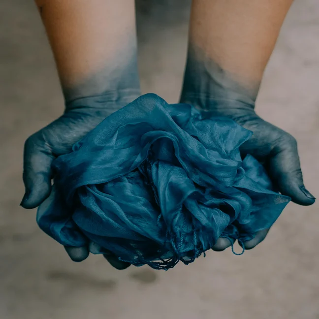 Blue-tinted fingertips gently hold blue fabric, set against a simple background, reflecting mindful craft and natural dye traditions.