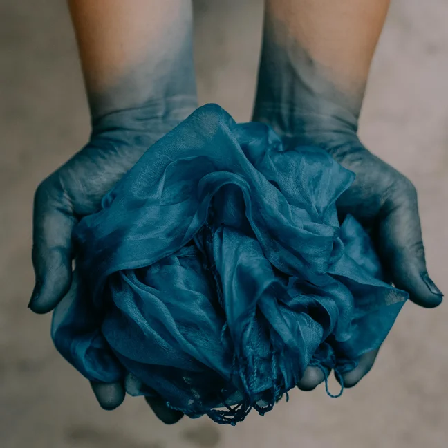 Blue-tinted fingertips gently hold blue fabric, set against a simple background, reflecting mindful craft and natural dye traditions.