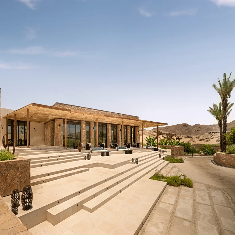 Contemporary structure with expansive windows and stone detailing, broad steps leading to shaded outdoor seating, surrounded by palms and desert views under clear blue skies.