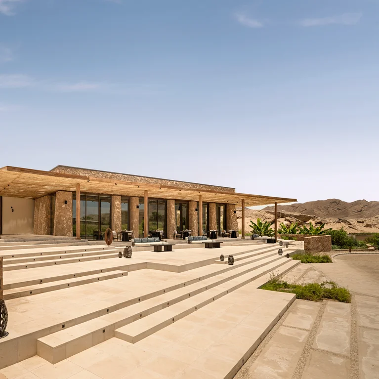 Contemporary structure with expansive windows and stone detailing, broad steps leading to shaded outdoor seating, surrounded by palms and desert views under clear blue skies.
