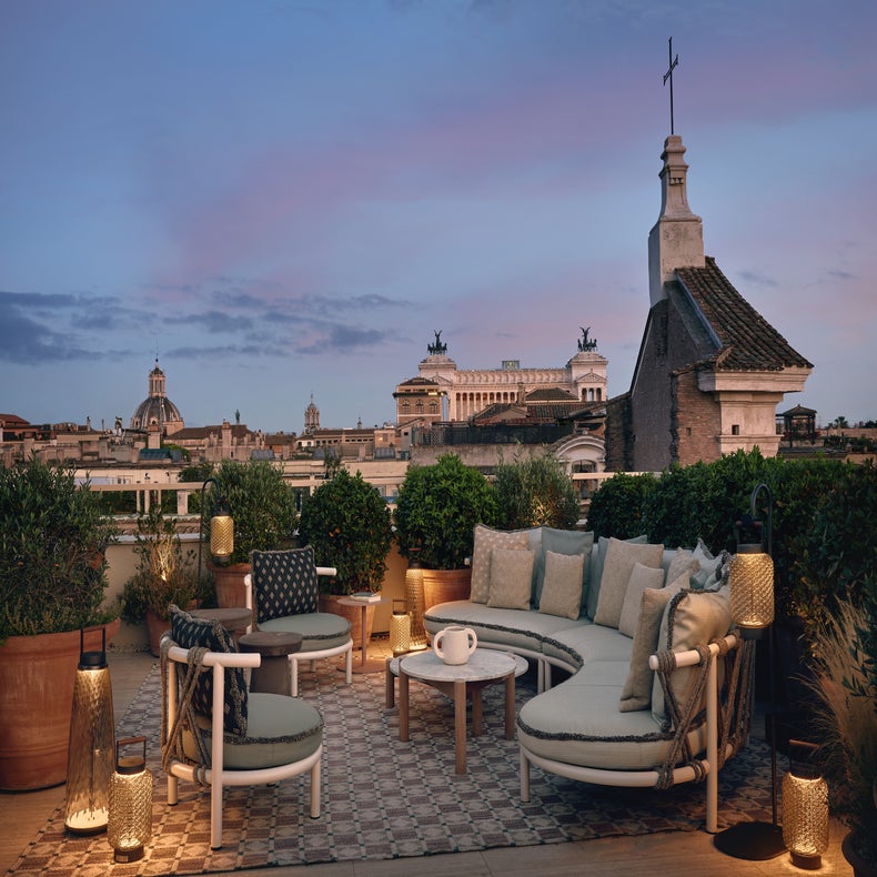 Terrasse avec des sièges confortables, une végétation abondante, un éclairage doux et des vues sur les bâtiments historiques et les dômes sous un ciel nocturne chaud.
