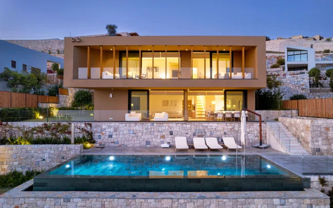 Sustainable villa featuring expansive windows, inviting interiors, outdoor seating, and an infinity pool on a stone terrace overlooking gentle hills at sunset.