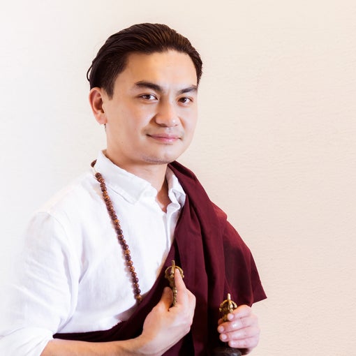 Individual in white shirt and maroon robe seated with a small bell and prayer beads, set against a neutral background, reflecting calm presence.