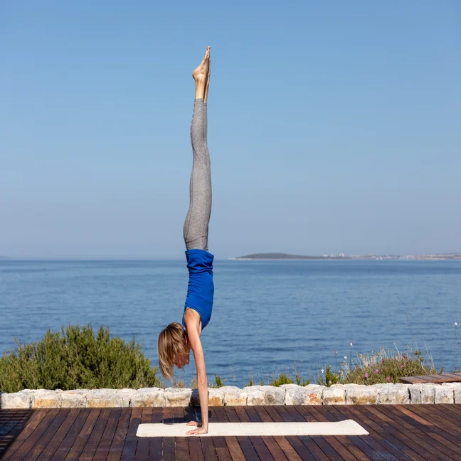 A guest practices a handstand on a yoga mat on a wooden deck with an ocean view.