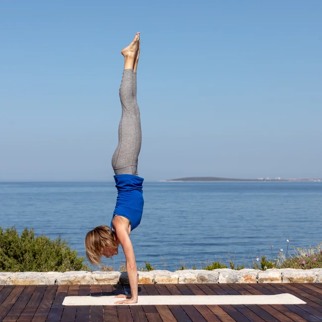 A guest practices a handstand on a yoga mat on a wooden deck with an ocean view.