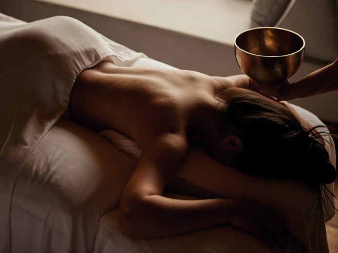 Guest on a massage table, draped in a white towel, with a practitioner holding a metal bowl near their back in a calm, softly lit treatment room.