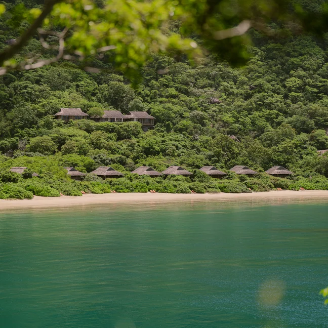 Sustainable pavilions set along a sandy shoreline, embraced by lush forest and overlooking calm turquoise waters.