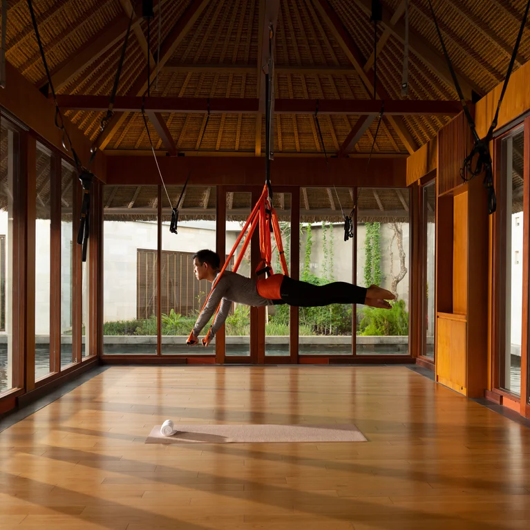 在明亮的工作室中,客人被悬挂在水平吊带上进行空中瑜伽,工作室铺有木地板、大窗户和天然茅草屋顶。.