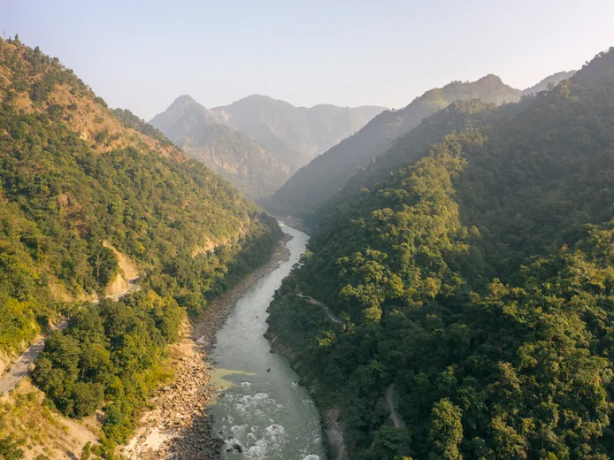 A clear river flows between green hills and mountains, with stone-lined banks and a dense forest canopy on both sides under an open sky.