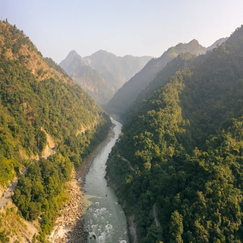 A clear river flows between green hills and mountains, with stone-lined banks and a dense forest canopy on both sides under an open sky.
