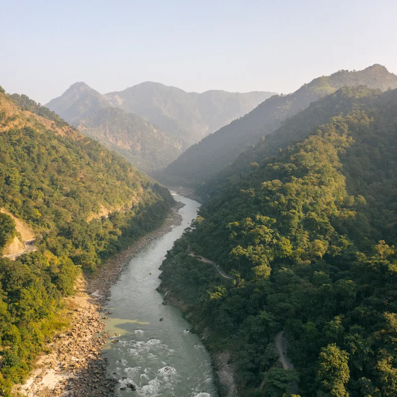 A clear river flows between green hills and mountains, with stone-lined banks and a dense forest canopy on both sides under an open sky.