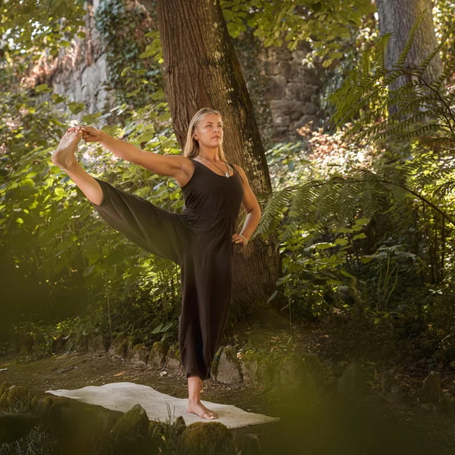 A practitioner in black yoga attire balances on one leg, extending the other upward on a mat set within a wooded garden.