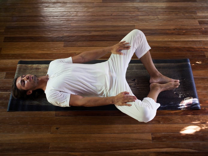Una invitada practica una postura de yoga reconstituyente sobre una esterilla en el interior, con las rodillas suavemente abiertas y las manos apoyadas en las piernas, sobre un suelo de madera lisa en un espacio tranquilo.