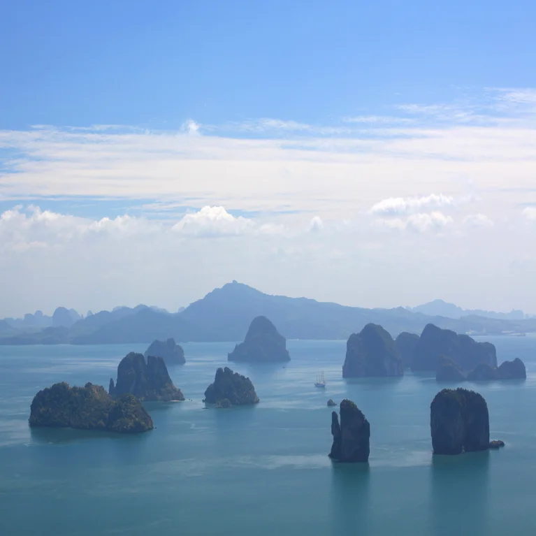 Islands with lush greenery emerge from tranquil blue waters beneath scattered clouds, while a distant shoreline of mountains stretches across the horizon.