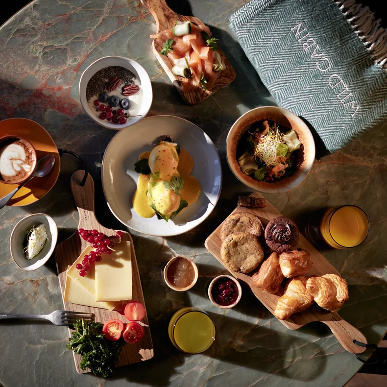 Table with coffee, juice, eggs Benedict, pastries, cheese, fruit, granola, avocado toast and a folded blanket with NUMBER CINQUE written on the label.