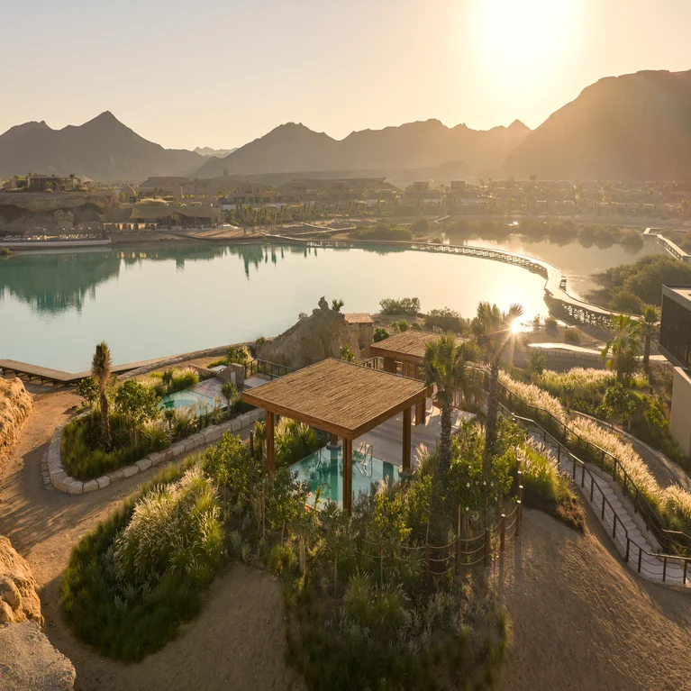 Wooden pergola beside a tranquil lake, surrounded by greenery and winding paths, with distant mountains across the water.