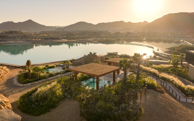 Wooden pergola beside a tranquil lake, surrounded by greenery and winding paths, with distant mountains across the water.