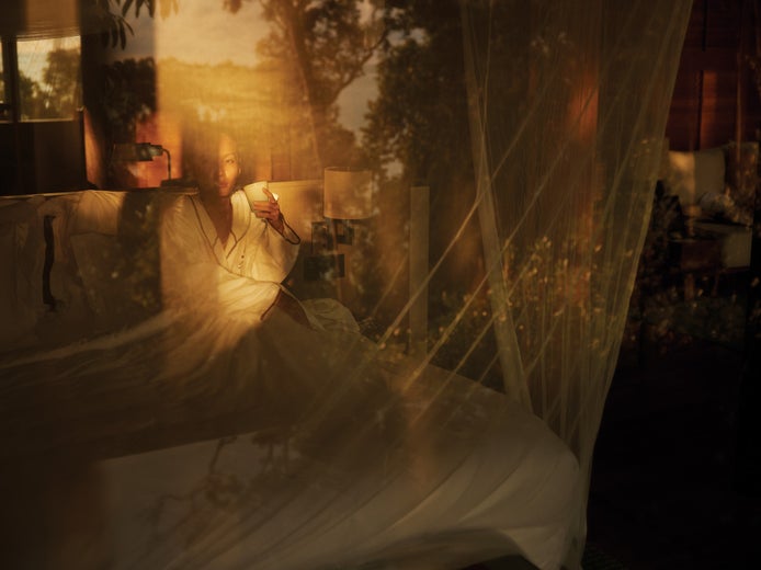 Guest in a white robe sits on a bed, enjoying a drink, with gentle sunlight and greenery reflecting on the nearby window.