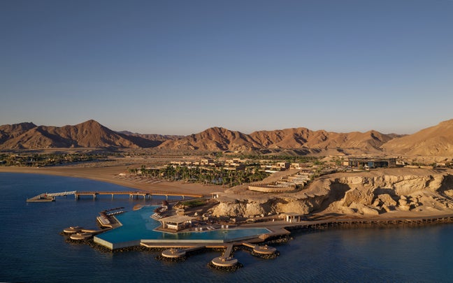 Pools and contemporary villas set beside the sea, framed by rugged mountains and desert terrain, featuring harmonious design within a natural coastal landscape.