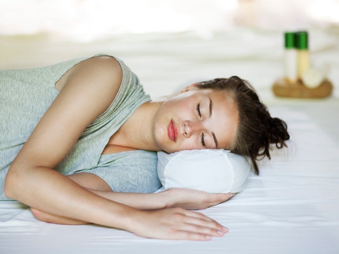 Una mujer de pelo castaño está tumbada de lado, apoyando la cabeza en una almohada sobre una cama blanca. Una suave luz natural entra en la serena habitación, creando un ambiente tranquilo.