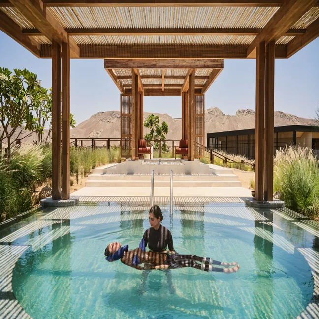 Two people enjoy a serene moment in a clear pool under a wooden pergola, with views of mountains and nearby resort buildings in the background.