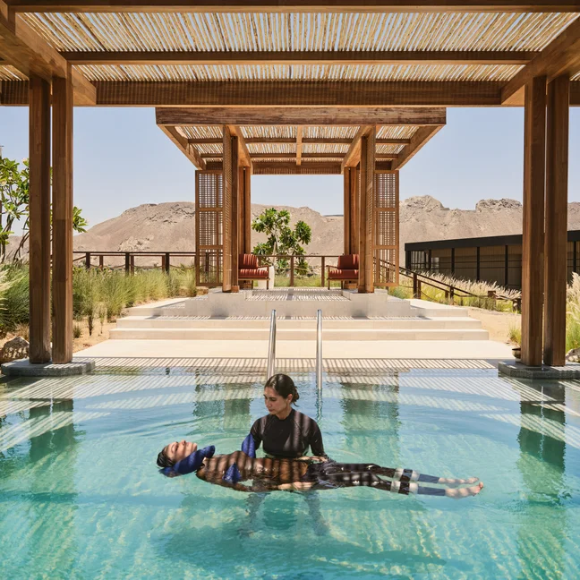 Two people enjoy a serene moment in a clear pool under a wooden pergola, with views of mountains and nearby resort buildings in the background.