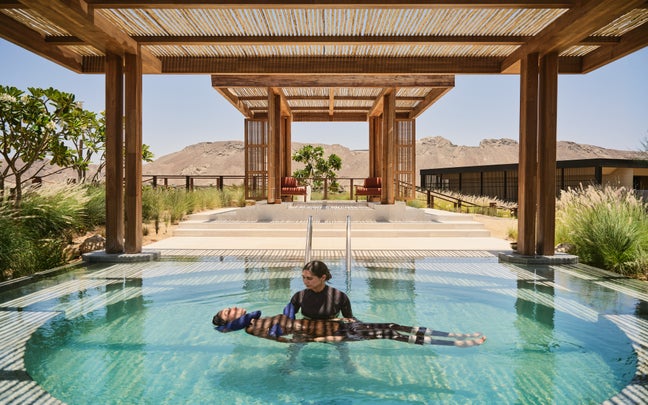 Two people enjoy a serene moment in a clear pool under a wooden pergola, with views of mountains and nearby resort buildings in the background.