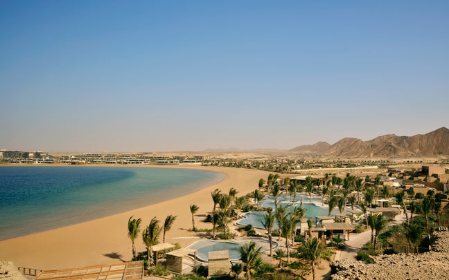 Sandy shoreline beside blue sea, with palm trees, pools, and thoughtfully designed resort buildings in front of mountains under clear sky.