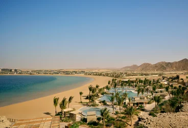 Sandy shoreline beside blue sea, with palm trees, pools, and thoughtfully designed resort buildings in front of mountains under clear sky.