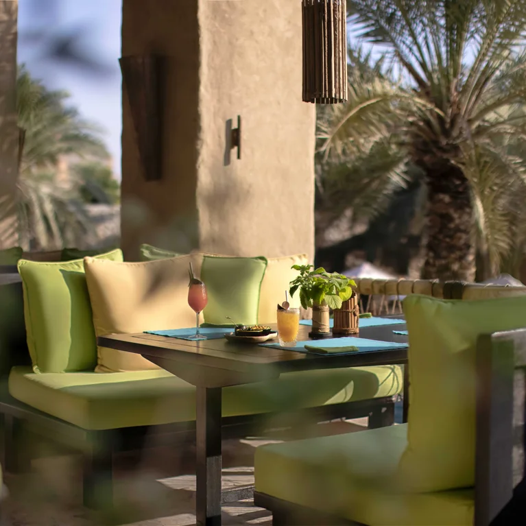 منطقة لتناول الطعام على الشرفة مع كراسي خضراء مبطنة وطاولة مجهزة لتناول الطعام، مظللة بأشجار النخيل والضوء الطبيعي.