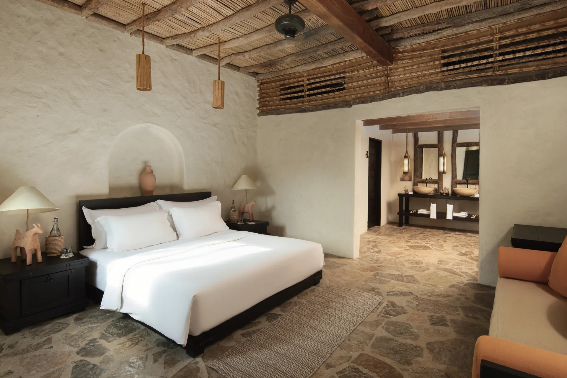 A pool villa bedroom under a ceiling of split cane laid over dark timber beams, white rendered walls with an arched niche holding a clay jar, a dark wood bed and a stone-flagged floor running through to the twin-basin bathroom.