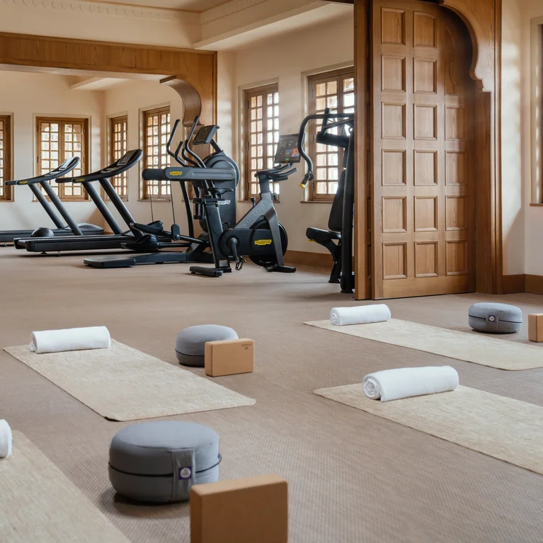 设备齐全的健康工作室配有跑步机、自行车、瑜伽道具和毛巾,自然光线充足,远处的墙壁上还有一位客人。.