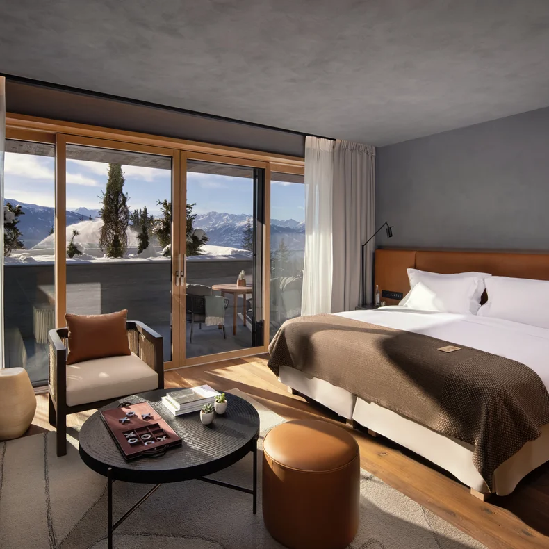 Spacious guest room featuring a king bed, sofa, and round table. Floor-to-ceiling windows open to a balcony with views of snow-covered mountains.