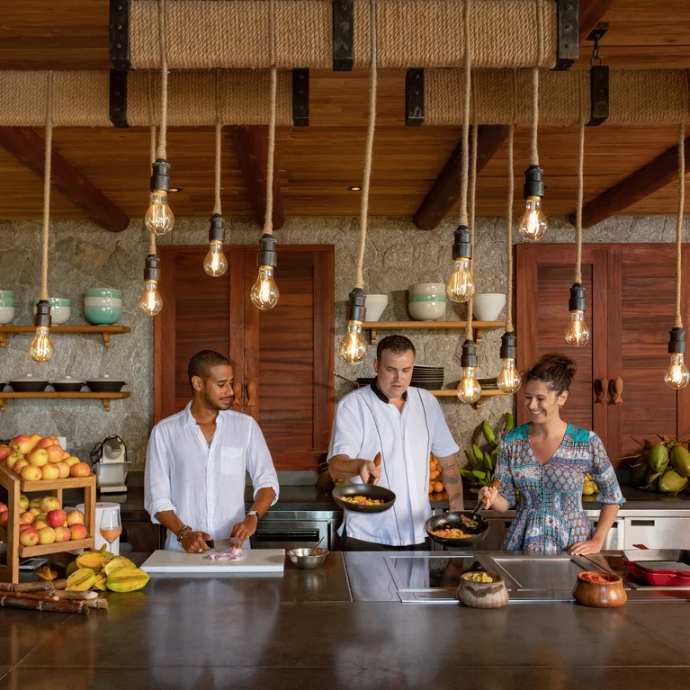 Los huéspedes preparan la comida en una cocina luminosa con estanterías de madera natural, lámparas colgantes, fruta fresca y cerámica bien ordenada.