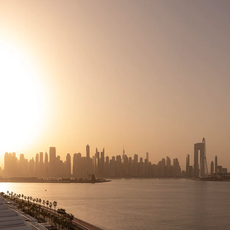 Dubai skyline with contemporary architecture, a prominent Ferris wheel, and tranquil water in the foreground on a hazy evening.