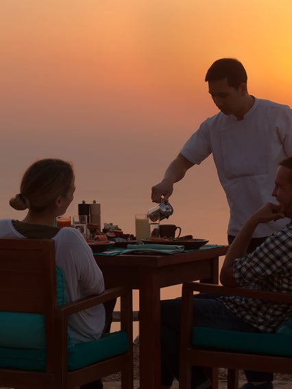 Un camarero lleva los platos a dos comensales que cenan al aire libre al atardecer, con vistas a un paisaje natural y una suave luz nocturna en el horizonte.