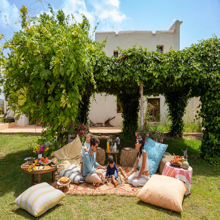 Three people sit together on a blanket with pillows and food in a sunlit garden, surrounded by greenery, outside a vine-covered white house.