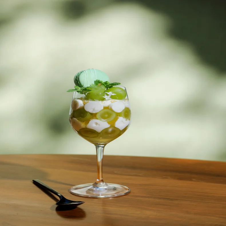 Glass dish with green grapes, plant-based cream, and a green macaron topped with mint; placed on a wooden table next to a dark spoon.