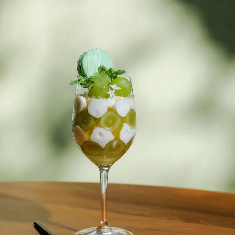 Glass dish with green grapes, plant-based cream, and a green macaron topped with mint; placed on a wooden table next to a dark spoon.